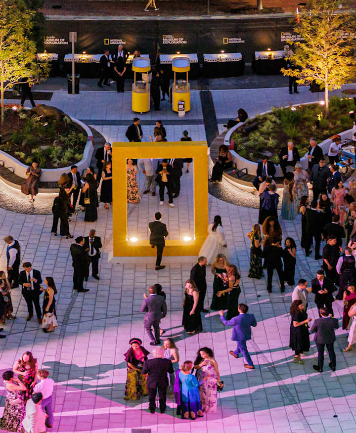 An aerial view of the Museum of Exploration's Courtyard at dusk shows a lively atmosphere with purple and pink accent lighting and large groups of people socializing throughout. 