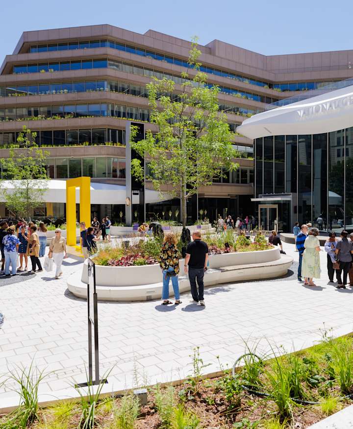 Visitors wander in the sunny outdoor courtyard of the Museum of Exploration, which features curved planters, a large yellow border icon, and the museum's glass front entrance.