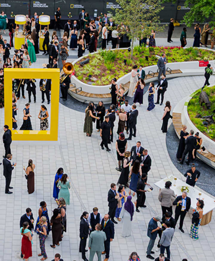 An overhead view shows a large crowd of guests in evening wear mingling in the outdoor courtyard of the Museum of Exploration, which features a spacious paved stone plaza, curved planters, and a large yellow border icon sculpture.