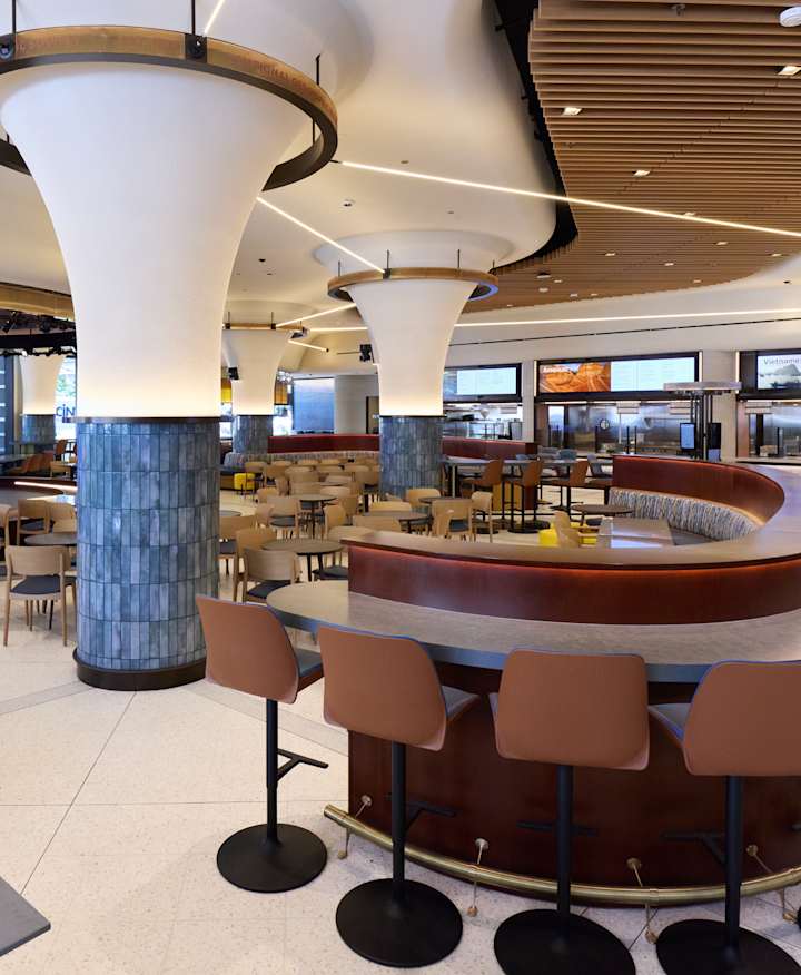The empty museum eatery featuring a large curved counter with stools and numerous dining tables, set against a background of flared pillars, food stations, and a small stage with a large digital screen displaying an expedition scene.