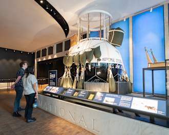 Two people stand side-by-side admiring a large round vessel that is wrapped with what appears to be large weighted bags. The vessel appears to be an object on display within an exhibition. 