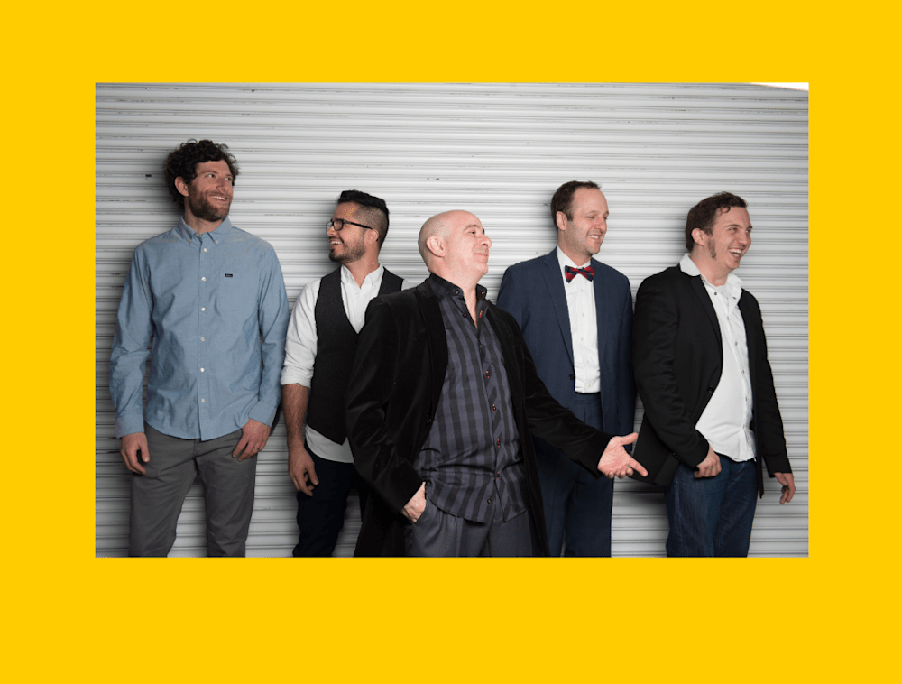 Five men standing side-by-side in front of a corrugated metal background. 