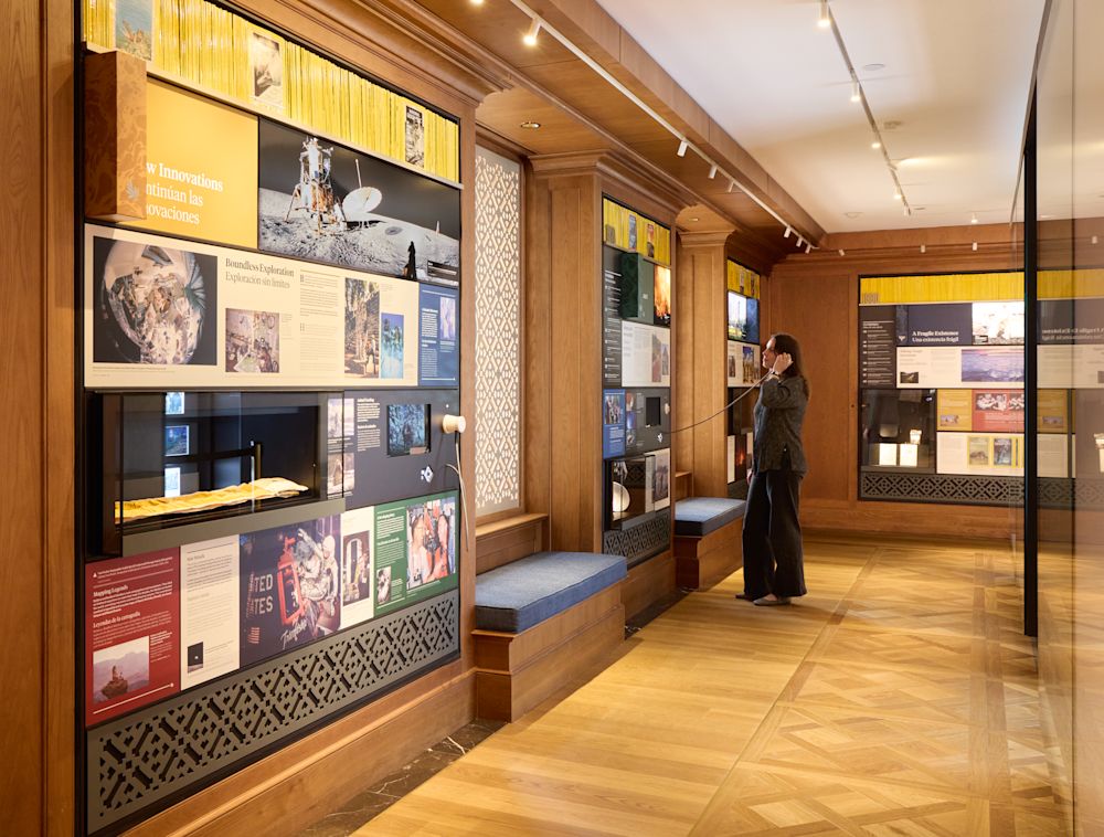 Una mujer de tez clara está de pie en un espacio expositivo del museo, sosteniéndose un auricular sobre la oreja. 