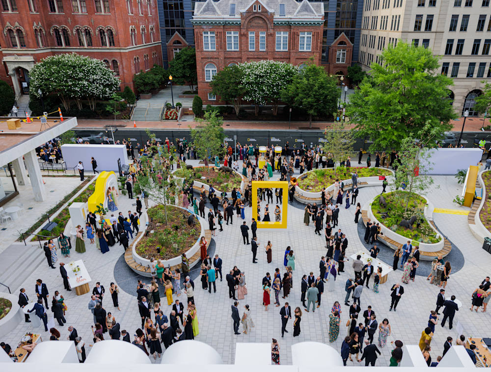 An overhead view shows a large crowd of guests in evening wear mingling in the outdoor courtyard of the Museum of Exploration, which features a spacious paved stone plaza, curved planters, and a large yellow border icon sculpture.