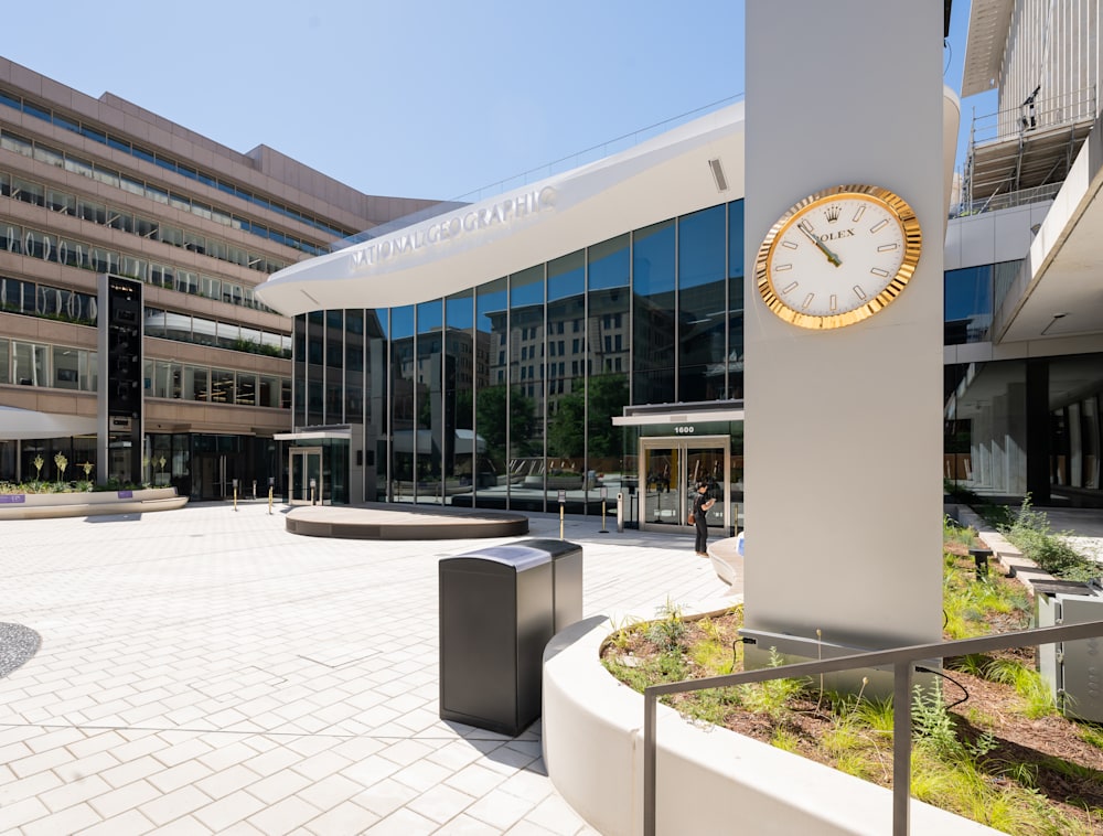 A sunny, empty outdoor courtyard at the Museum of Exploration featuring a spacious paved stone plaza, a low oval stage, and a large gold Rolex clock mounted on a pillar in front of the museum's glass facade. 