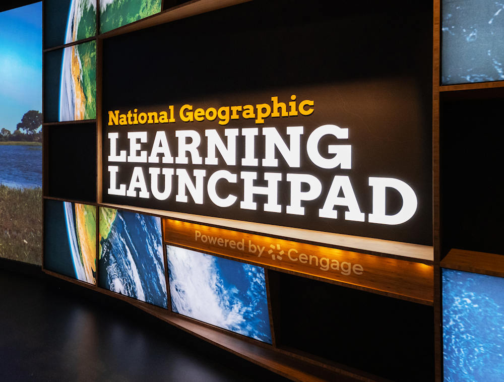 A round, globe-like multimedia display with a central sign reading "National Geographic Learning Launch Pad Powered by Cengage" framed by images of Earth, next to a large curved panoramic screen showing a wetland landscape.