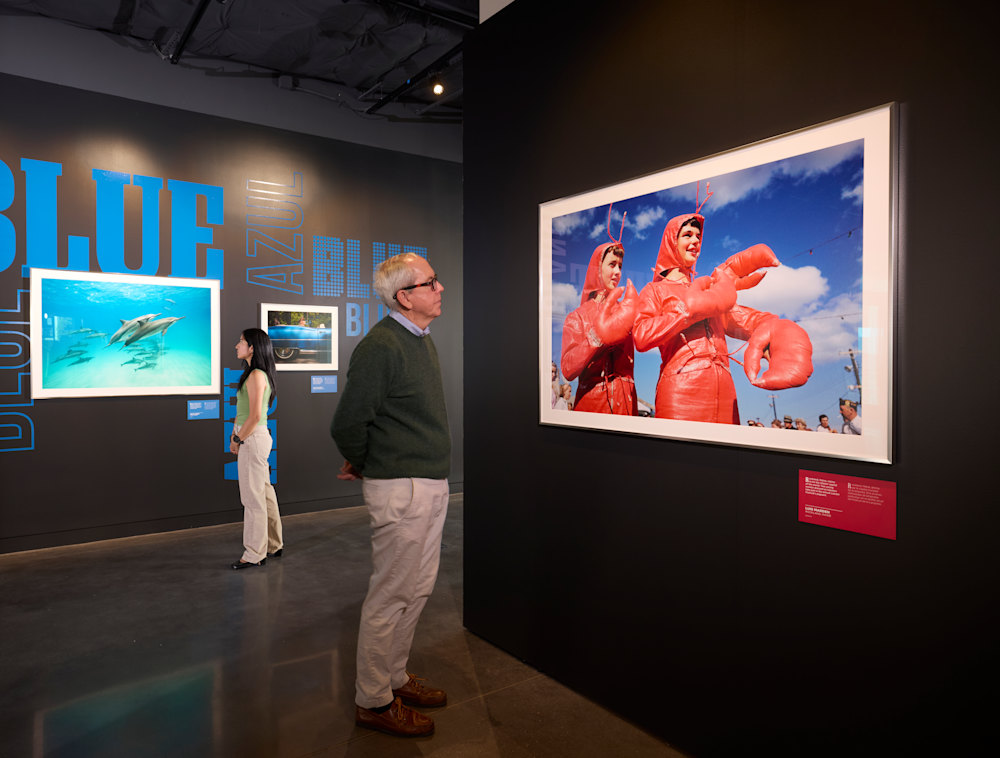 Two individuals study framed photography inside the Red, White & Blue exhibition.