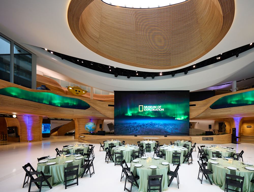 An expansive view of the Pavilion. Its wide space is filled with round, decorated tables. Screens throughout the space show the Northern Lights. The walls are of a curving, light wood and the floors white marble.