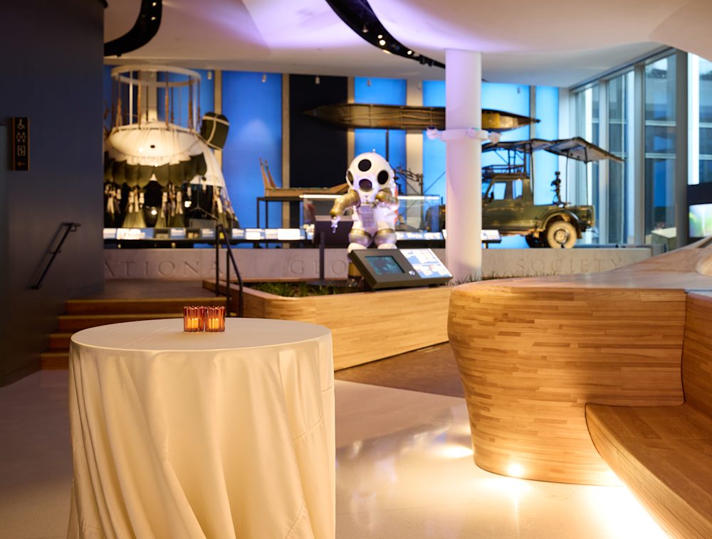 An exhibit space with a long wooden bench and marble floors, with a tall decorated table in the foreground. In the background are large displays, including a balloon gondola and a diving suit.