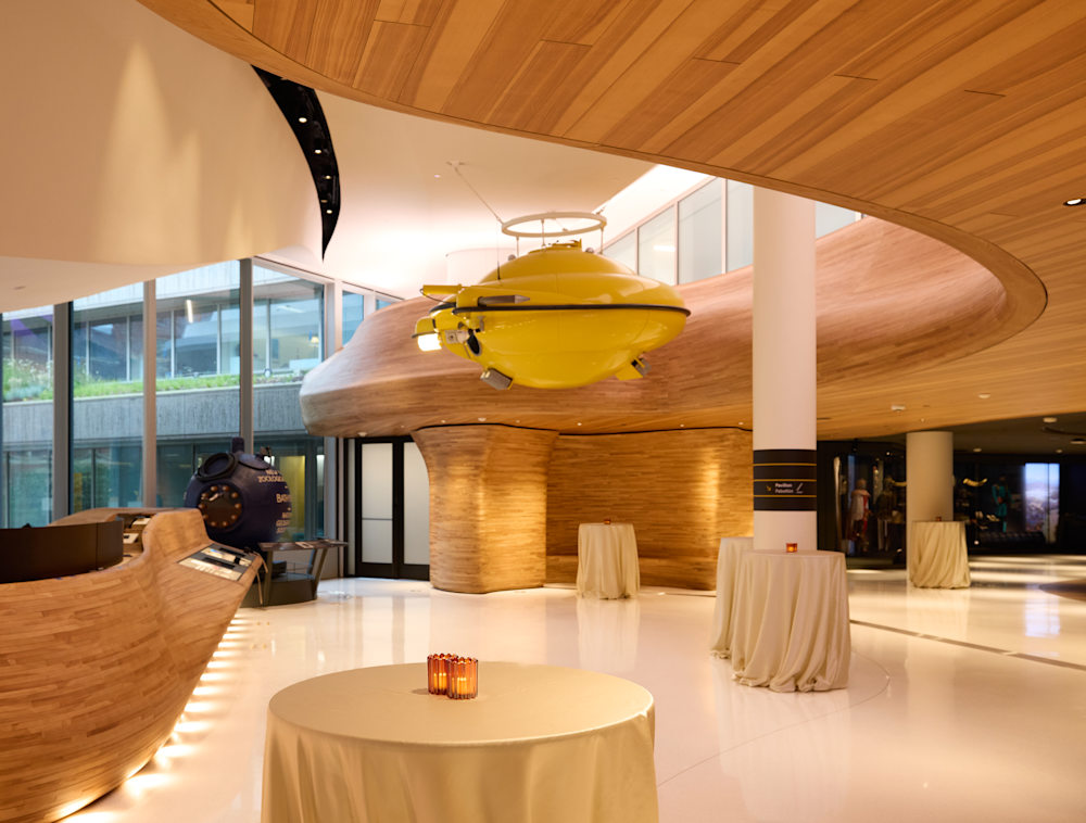 A small yellow submersible hangs from the ceiling of an exhibit space. The walls are a light-colored wood. Tall, decorated tables dot the space.