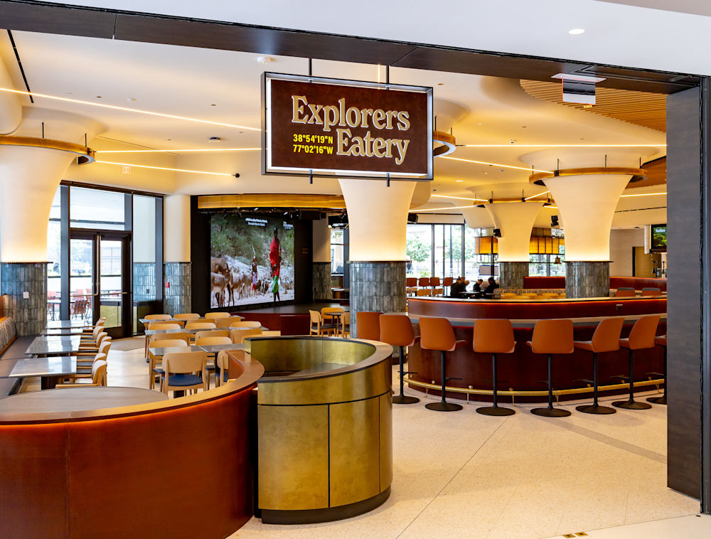 A hanging "Explorers Eatery" sign marks the entrance to the museum's cafe, revealing a spacious interior with tables, booth seating, a curved counter, flared pillars, and a small stage.