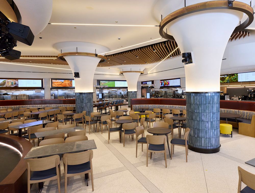 The empty museum eatery featuring an open dining area filled with numerous tables and chairs, framed by large flared pillars, slatted wood ceiling designs, and a line of food stations in the background.