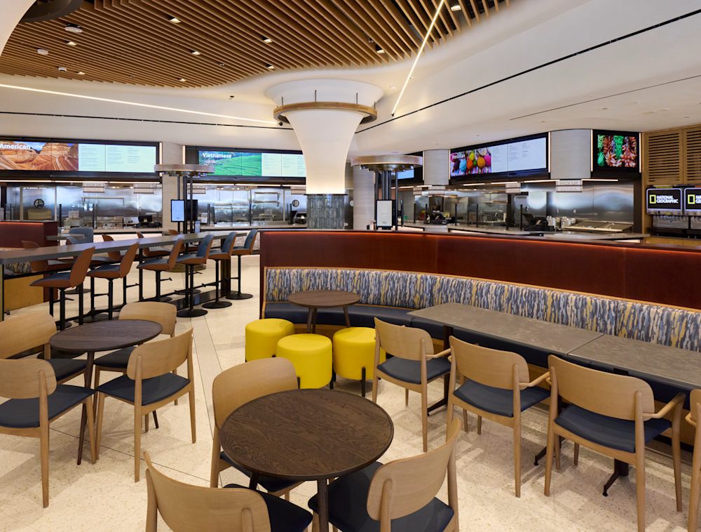 The empty museum eatery featuring a variety of seating options in the foreground, including dining tables with wooden chairs, bright yellow ottomans, and a curved booth, with food stations and digital menu boards lining the background.