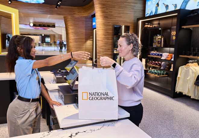 A woman grabs a retail bag from a cashier. The bag is white and has the National Geographic logo printed on it. 