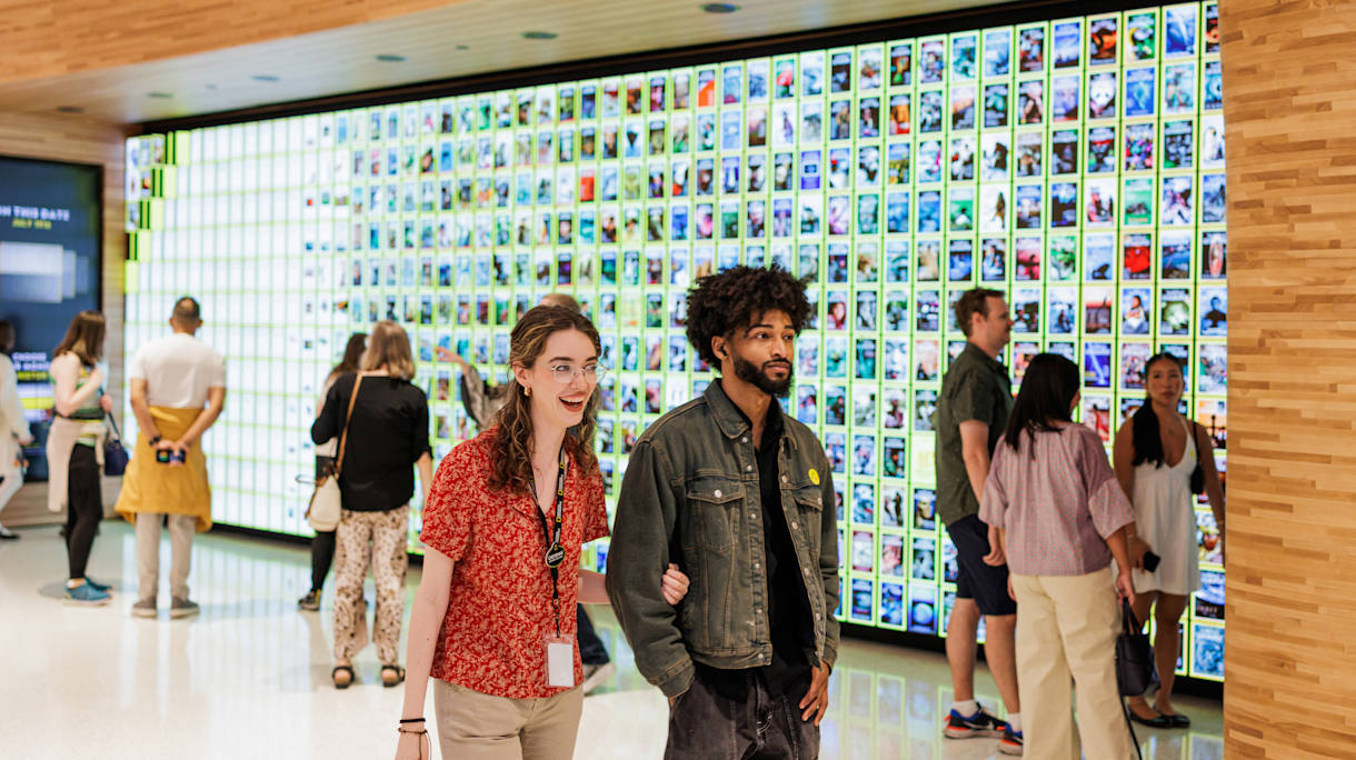 A woman with light skin tone and a man with dark skin tone walk arm-in-arm past a large media wall displaying National Geographic magazine covers.