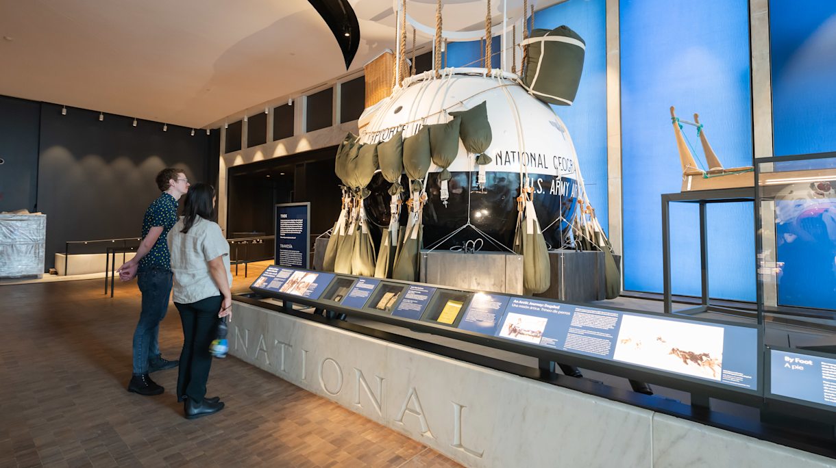 Two people stand in front of a large blue-and-white balloon gondola on display in an exhibit gallery.