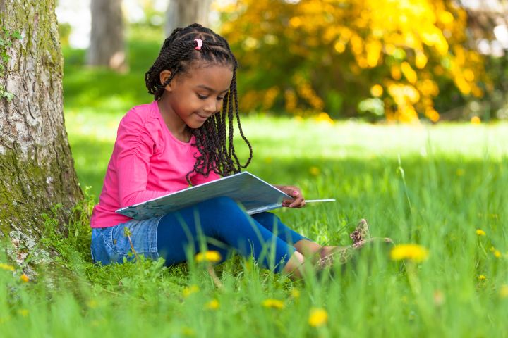 Outdoor portrait of a cute young black little girl reading a book - African people