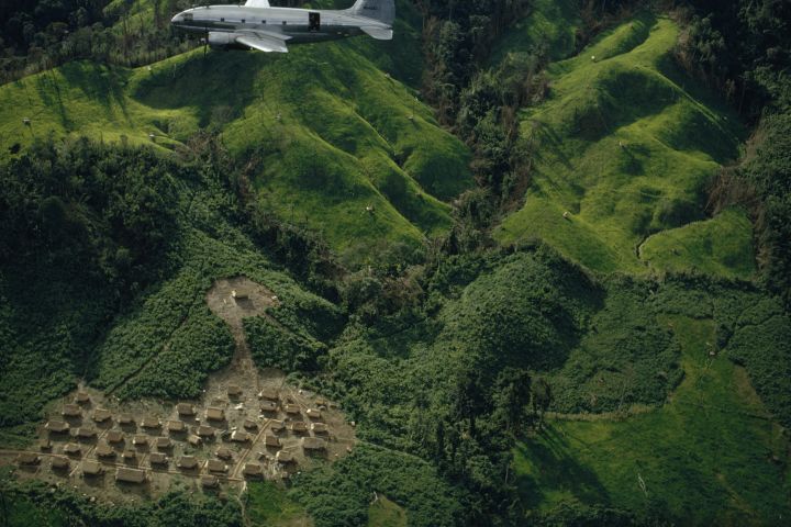 Airplane drops rice and corn above refugee village amid rugged hills.