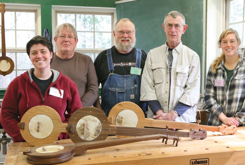 Photo of people standing behind a woodworking bench with unfinished banjos.