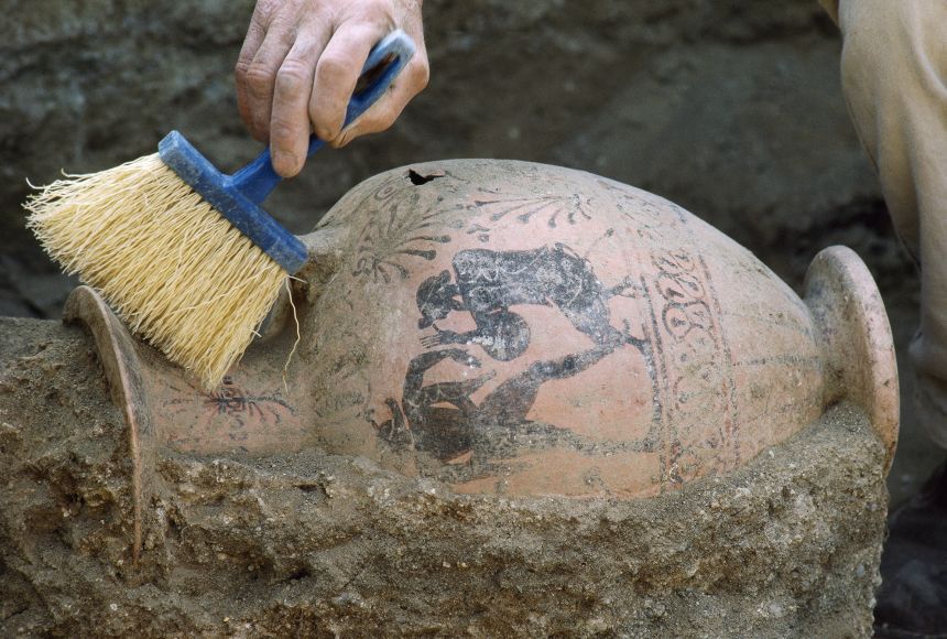 Archaeologists carefully excavate an ancient vase from hard-packed soil with soft brushes.
