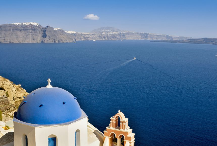 An image of a tiny boat sailing away from the coast with a domed building in the lefthand forgroud and sharply rising cliffs in the background.